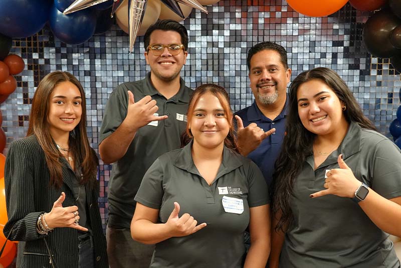 Group of attendees gather indoors during a UTEP Engineering homecoming event while taking a selfie, with several participants making hand gesture signs and standing together in front of a decorative event backdrop and balloon display.